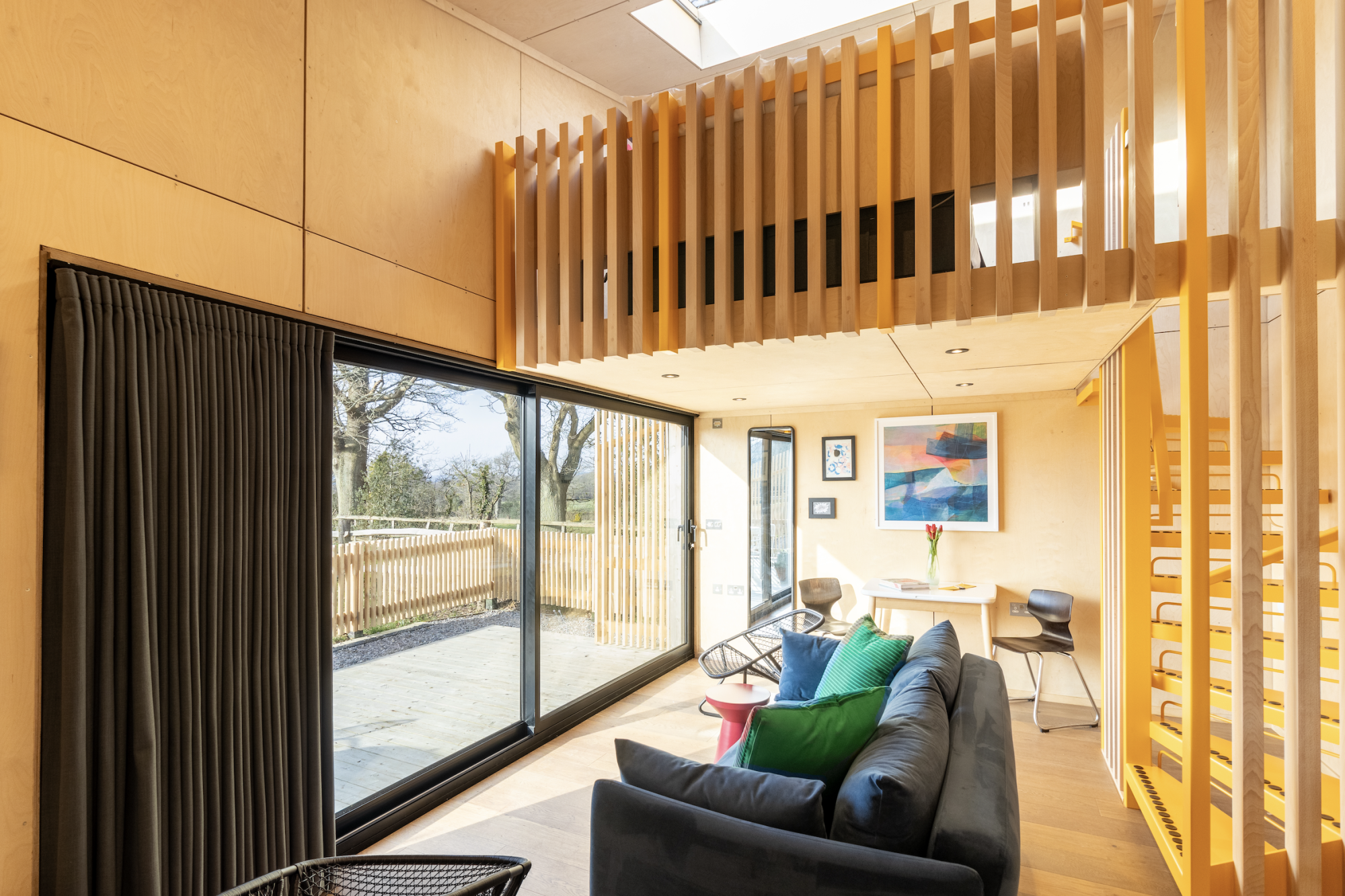Interior of a modern timber lodge featuring a plywood-lined living room, a yellow mezzanine sleeping loft with timber slats, and large sliding glass doors opening to a deck.